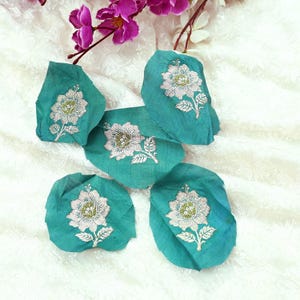 Peut inclure: Cinq pièces de tissu turquoise avec des motifs floraux brodés. Chaque pièce présente une fleur blanche et verte avec des coutures détaillées. Les pièces sont disposées sur une surface blanche texturée, avec des fleurs violettes en arrière-plan.