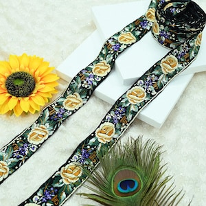 Peut inclure: Ruban noir avec broderie de roses jaunes, feuilles vertes et fleurs violettes. Le ruban est enroulé et posé à plat. Un tournesol jaune et une plume de paon sont également dans l'image.