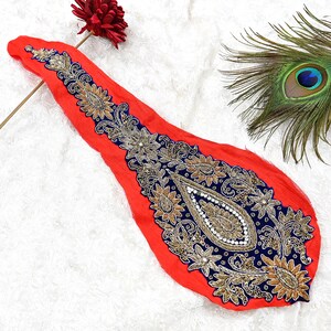 May include: A red fabric applique with detailed gold, navy blue, and white embroidery. The design incorporates floral and paisley motifs. A peacock feather and a red flower are visible in the background.