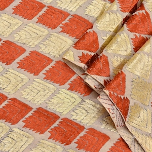 May include: A fabric featuring a repeating geometric pattern of alternating orange and metallic gold chevron-style leaf shapes arranged in diagonal rows on a light beige background, with the material folded to show texture and depth.