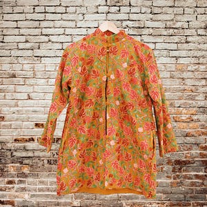 May include: Mustard-colored long-sleeve jacket with a floral pattern in red and green. The jacket has a mandarin collar and is buttoned down the front. It is hanging on a wooden hanger against a brick wall.