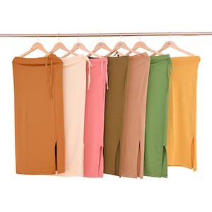 Puede incluir: Una fila de siete faldas largas en varios colores, incluyendo mostaza, verde oliva y rosa rubor, cuelgan de perchas de madera. Cada falda tiene una abertura lateral y un cordón en la cintura. Las faldas están hechas de tela suave.