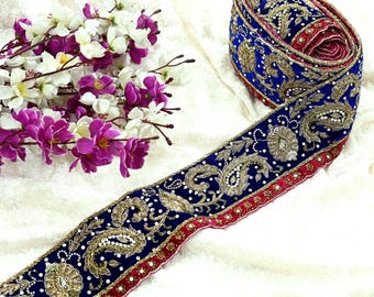 May include: A rolled blue and gold embroidered trim with a red border. The trim features intricate gold floral and paisley designs with white bead accents. A bundle of purple and white flowers is in the background.