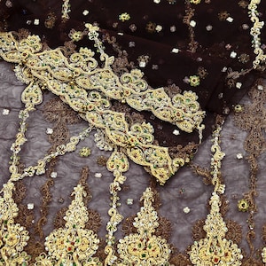 Op de afbeelding: Close-up van een bruine stof met ingewikkelde gouden borduursels. Het ontwerp heeft sierlijke bloempatronen, versierd met kleurrijke kralen. De randen zijn afgewerkt met een gedetailleerde gouden rand, wat een luxueuze en elegante uitstraling creëert.