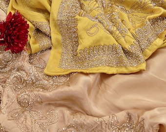 May include: A close-up of a yellow and beige textile with intricate silver embroidery. The yellow fabric is folded, revealing detailed floral patterns and beaded edges. A deep red flower is visible in the upper left corner, adding a pop of color.