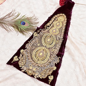 Könnte beinhalten: Ein tief burgunderfarbenes Samttextil mit aufwendiger Goldstickerei. Das Design zeigt große, verzierte Blumen- und Paisley-Muster. Eine Pfauenfeder liegt daneben und fügt einen Hauch von Farbe und Textur hinzu. Das Textil ist wahrscheinlich ein dekoratives Stück oder Teil eines Kleidungsstücks.