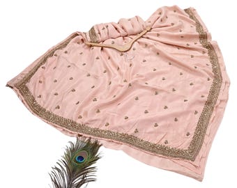 May include: A light pink sari with intricate gold embroidery along the border and scattered across the fabric. The sari is draped over a wooden hanger. A peacock feather is placed near the sari.