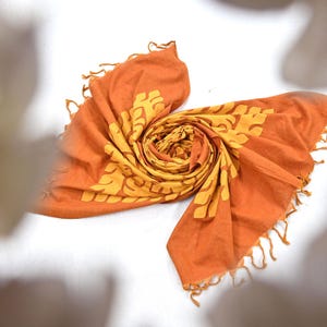 May include: An orange scarf with a yellow floral pattern and fringed edges. The scarf is rolled up in a spiral shape, showcasing the design. The scarf is made of a soft material.