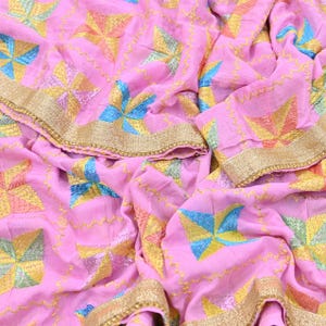 Könnte beinhalten: Ein hellrosa Stoff mit einem sich wiederholenden Muster aus vierzackigen Sternen in Blau-, Gelb- und Grüntönen. Das Textil ist mit goldfarbenen Metallfadenstickereien akzentuiert und mit einer breiten goldfarbenen Metallborte versehen.