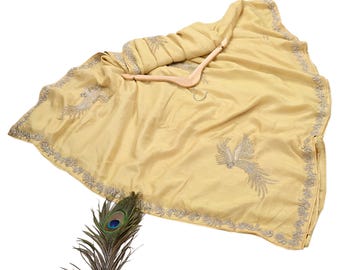 May include: A pale yellow sari with intricate silver embroidery along the borders and a detailed bird design. The fabric is draped over a wooden hanger. A peacock feather is placed near the sari.