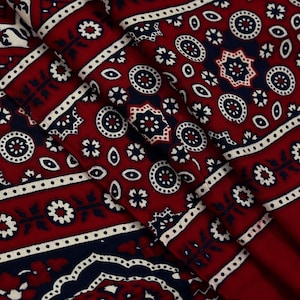Puede incluir: Primer plano de una tela doblada con un patrón repetitivo. El diseño presenta colores blanco, azul marino y rojo-burdeos. El patrón incluye formas florales y geométricas, como círculos, estrellas y puntos. La tela parece ser de algodón o un material similar.