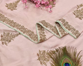 May include: A light pink fabric with gold floral embroidery and a decorative ribbon with a light green border. Pink flowers and a peacock feather are arranged on the fabric.