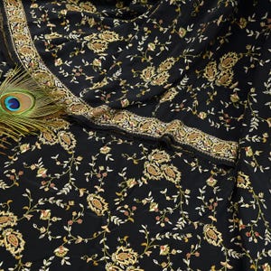 Puede incluir: Tela negra con un intrincado estampado floral en oro, crema y rojo. La tela tiene un borde bordado detallado. Una pluma de pavo real es visible en la esquina, agregando un toque de elegancia.