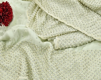 May include: A light green fabric with intricate pearl and sequin embellishments. The fabric is draped, with a section resembling a jacket or top, and a red flower in the upper left corner. The design is detailed and elegant.