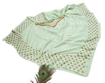 May include: A light green saree with a decorative border of small, square, gold-colored embellishments. The saree is draped over a wooden hanger. A peacock feather is in the foreground.