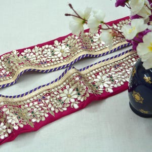 Puede incluir: Borde de tela rosa con adornos dorados y plateados, con diseños de flores blancas y un borde azul.