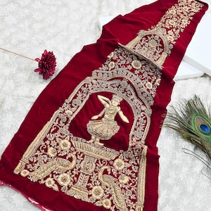 Puede incluir: Textil rectangular largo de terciopelo rojo oscuro con intrincados bordados dorados. El diseño presenta un arco central con una figura de bailarina, motivos florales y pavos reales. El textil es probablemente una pieza decorativa.