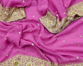 May include: A vibrant pink fabric with intricate gold embroidery and embellishments. The fabric is draped, showcasing the detailed patterns along the borders and scattered across the surface. The gold accents provide a striking contrast to the pink.