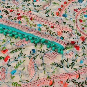 Könnte beinhalten: Ein weißer Stoff mit aufwendigen Stickereien in roten, grünen und blauen Blumen- und Rankenmustern. Das Textil ist an der Kante mit einem leuchtend türkisfarbenen Band verziert, das mit kleinen, runden, passenden türkisfarbenen Pompons besetzt ist.