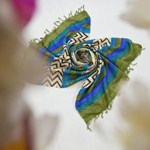 May include: A rolled scarf with a mix of blue, green, and cream colors. The scarf features a chevron pattern and stripes, with fringed edges. The scarf is made of a lightweight fabric, perfect for adding a touch of style.