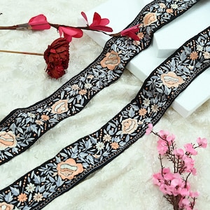 Può includere: Due nastri neri ricamati con motivi floreali. I nastri presentano rose color pesca, foglie bianche e grigie e piccoli fiori bianchi. I nastri sono esposti su una superficie bianca con fiori rosa.