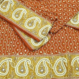 May include: An orange textile with intricate gold paisley and sequin detailing. The fabric features a repeating pattern of small, gold sequins and larger, ornate paisley borders. The design is likely for a garment or decorative item.