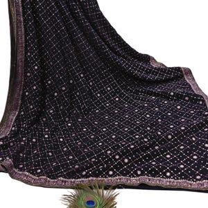 May include: A black fabric with a grid pattern of small, light-colored dots and floral accents. The border features a decorative trim with gold and pink accents. A peacock feather is in the foreground.