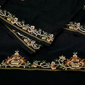 May include: A black fabric garment featuring intricate embroidery along the hem. The embroidery displays a floral and paisley pattern in shades of rust, gold, cream, and olive green, accented by thin, parallel gold-colored stitched lines.