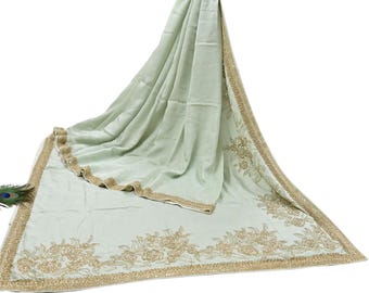 May include: A light green sari with intricate gold floral embroidery along the borders and edges. The fabric drapes elegantly, showcasing the detailed craftsmanship. A peacock feather is visible in the corner. The sari is a traditional garment.
