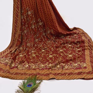 May include: A draped, rust-colored textile with intricate gold embroidery featuring floral patterns. The fabric has a textured, dotted design and a decorative border. A peacock feather rests in the foreground.