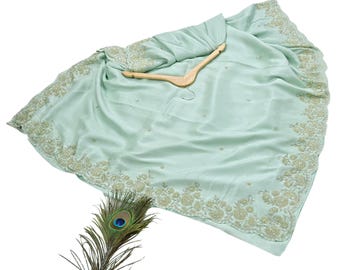 May include: A light green saree with intricate gold embroidery along the borders and scattered across the fabric. The saree is draped over a wooden hanger. A peacock feather is placed near the saree.