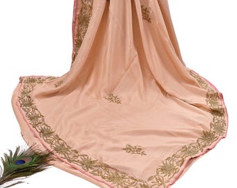 May include: A peach-colored sari with gold embroidery along the border and scattered across the fabric. The sari has a pink trim and is draped, showcasing the intricate design and texture. A peacock feather is visible in the lower left corner.