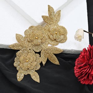 Peut inclure: Une applique florale en perles dorées composée de trois fleurs superposées et de plusieurs feuilles pointues. Le motif est réalisé avec des perles de rocaille dorées métallisées et présente une petite perle dorée lisse au centre de chaque fleur.