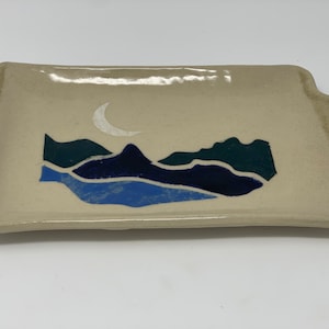 Op de afbeelding: Een keramische serveerschaal met een crèmekleurige achtergrond en een blauwe, groene en witte berg scène met een halve maan.