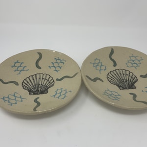 May include: Two ceramic plates with a beige background and blue and green designs. Each plate features a black and white seashell illustration and a net pattern.