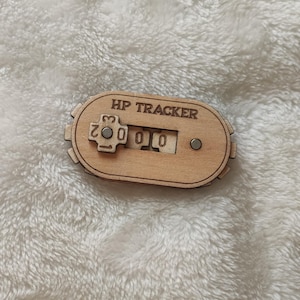 Op de afbeelding: Een houten HP-tracker met een wijzerplaat die kan worden gedraaid om van 0 tot 20 te tellen. De tracker is gelabeld met "HP TRACKER".