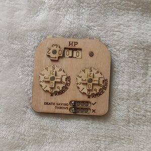 Könnte beinhalten: Holz-Charakter-Tracker mit Zifferblättern für Trefferpunkte, Rüstungsklasse und Angriffsbonus. Es hat auch einen Bereich für Todesrettungswürfe mit Häkchen und Kreuzen.
