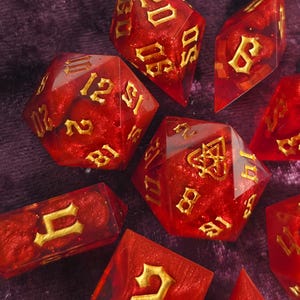 Pode incluir: Um conjunto de dados poliédricos vermelhos e dourados, perfeitos para jogos de tabuleiro. Os dados são transparentes com números e símbolos dourados. O conjunto inclui várias formas, como D20, D6 e D4, todos com um efeito brilhante.