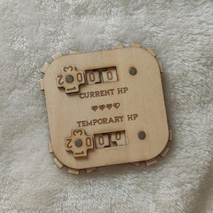 Può includere: Contatore in legno con quadranti per tenere traccia dei punti ferita attuali e temporanei. I quadranti sono impostati su 000 e 000. Il testo "CURRENT HP" e "TEMPORARY HP" è stampato sul contatore.