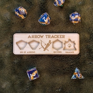 Pode incluir: Rastreador de flechas de madeira com ranhuras para o número de flechas, aljavas e danos. O rastreador é feito de madeira marrom claro e tem um acabamento de madeira natural. Há cinco dados na imagem, quatro dos quais são translúcidos com acentos azuis e dourados, e um é opaco com acentos dourados.