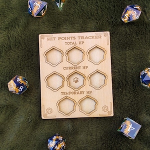 Op de afbeelding: Een houten hitpoints-tracker met negen slots voor dobbelstenen. De tracker heeft de woorden "Hit Points Tracker", "Total HP", "Current HP" en "Temporary HP" erop gedrukt. De tracker is omringd door blauwe en gouden dobbelstenen.