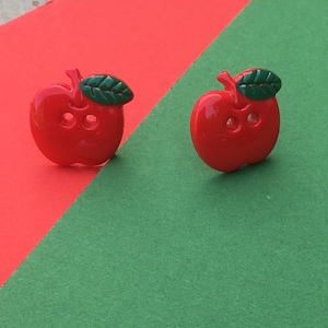 Pode incluir: Dois botões brilhantes em forma de maçã vermelha, cada um com uma pequena folha verde presa ao caule. Os botões são projetados com dois furos no centro para costura, apresentando um acabamento liso e brilhante e uma cor vermelha sólida vibrante.