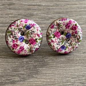May include: A pair of white ceramic button earrings with a floral pattern in shades of pink, purple, and green.