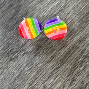 May include: A pair of round stud earrings shaped like buttons, each featuring a vibrant rainbow striped pattern with red, orange, yellow, green, blue, and purple colors. The earrings have a smooth, glossy finish and clear plastic earring posts.