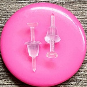 Puede incluir: Dos respaldos de pendientes de plástico transparente sobre un fondo rosa. Los respaldos están diseñados para ser usados con pendientes de botón.
