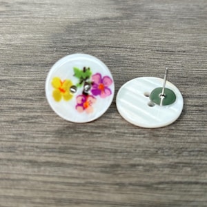 May include: A pair of white shell earrings with a floral design. The front of the earrings feature a yellow, pink, and purple flower design. The back of the earrings are plain white with a silver post.