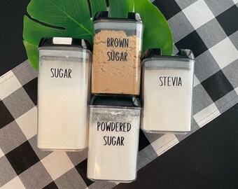May include: Four clear, rectangular food storage containers with black lids, labeled "SUGAR", "BROWN SUGAR", "STEVIA", and "POWDERED SUGAR". The containers are arranged on a black and white checkered table runner, with a green leaf in the background.