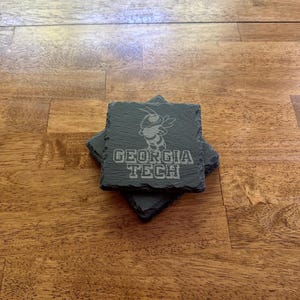 May include: A stack of four square, dark gray slate coasters. Each coaster features an etched image of a yellow jacket mascot and the words "GEORGIA TECH" in white lettering. The coasters are arranged on a wooden surface.