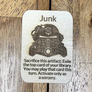 Könnte beinhalten: Eine Holzkarten mit einer Schwarzweißillustration eines Roboterkopfes. Die Karte trägt die Aufschrift "Junk" und den Text "Opfere dieses Artefakt: Verbann die oberste Karte deiner Bibliothek. Du kannst diese Karte in diesem Zug spielen. Aktiviere nur als Sorcery."