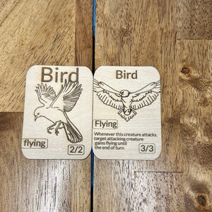 Könnte beinhalten: Zwei Holzkarten mit Illustrationen von Vögeln. Die Karte links zeigt einen Vogel mit dem Text "flying" und "2/2". Die Karte rechts zeigt einen Vogel mit dem Text "Flying" und "Whenever this creature attacks, target attacking creature gains flying until the end of turn" und "3/3".
